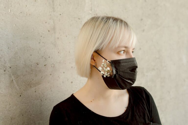 Blonde woman wearing a black face mask adorned with white flowers against a concrete wall.